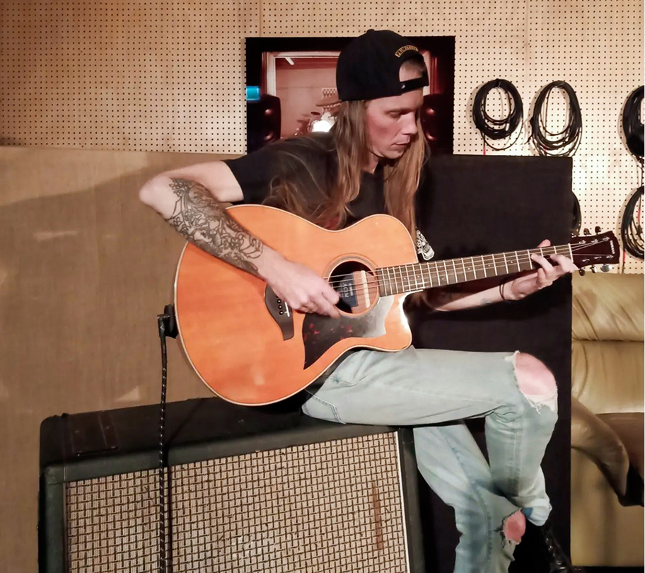 Man with long hair and a hat plays an acoustic guitar in a music studio located in Fairbridge Village
