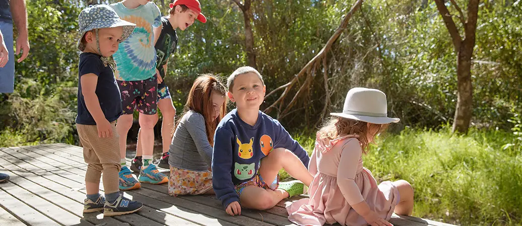 A group of kids sits on a wooden path, looking for a fish in a pond and enjoying nature together at Fairbridge Village
